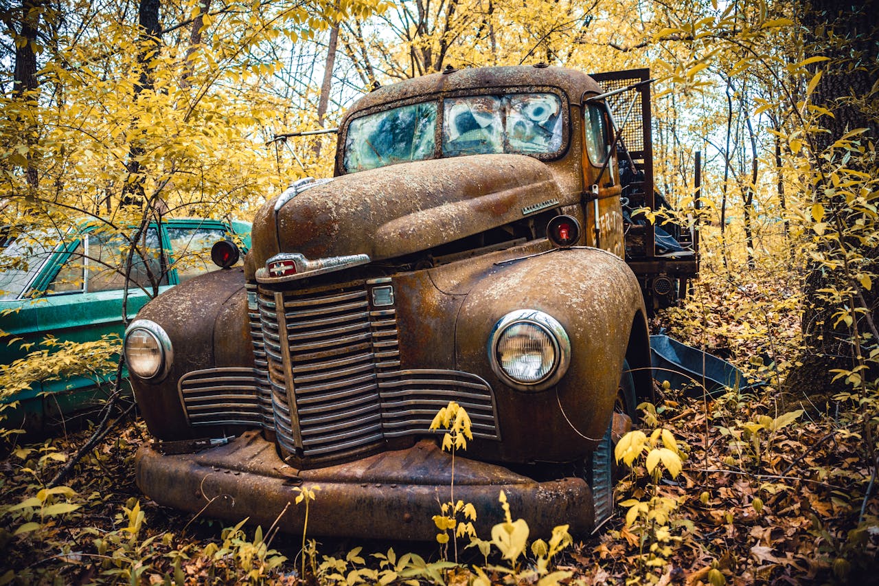 A vintage, rusty truck sits abandoned in a vibrant autumn forest, surrounded by yellow leaves.