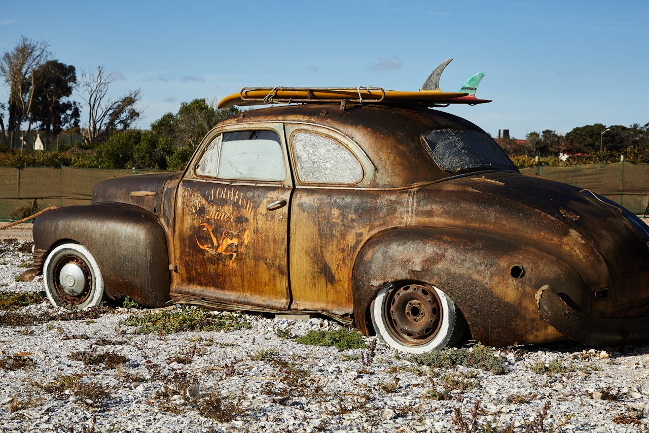 A weathered vintage car with surfboards on top, surrounded by a desolate landscape.